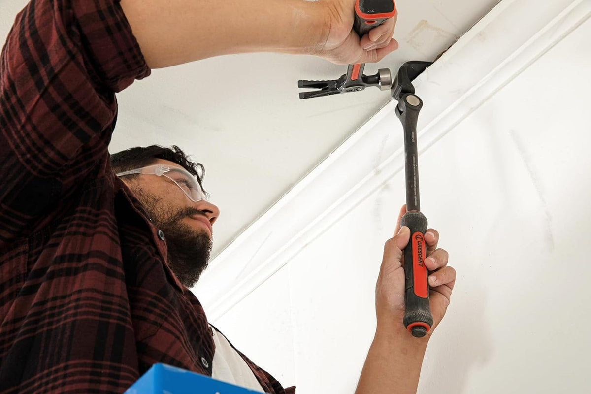 A worker in a plaid shirt and safety glasses uses the Crescent indexing pry bar to remove crown molding, while tapping the bar's striking surface with a hammer.