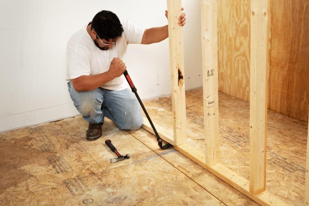 A man kneeling on a job site using the high-leverage Crescent 30-inch indexing flat pry bar to dismantle wooden wall framing.