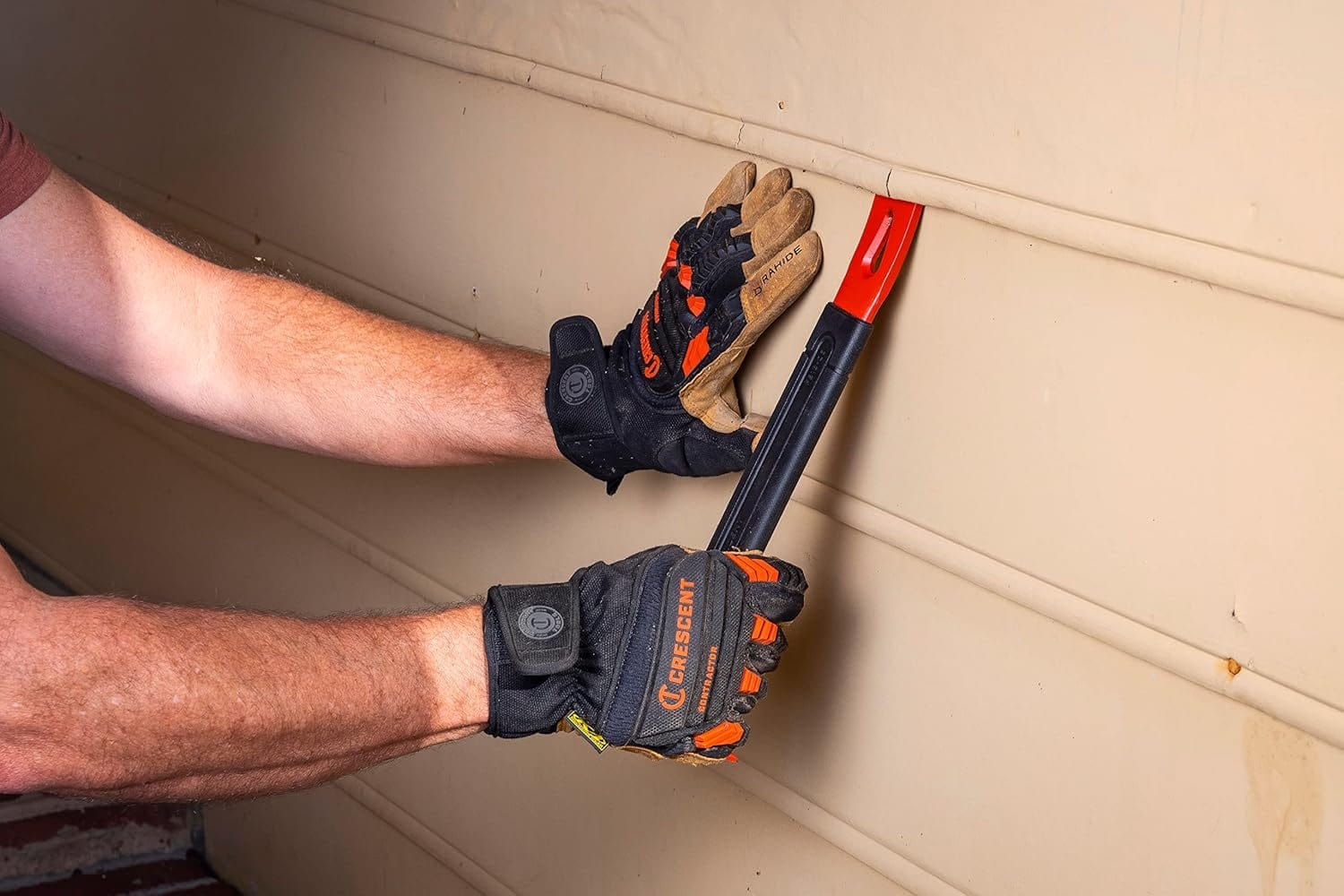Action shot of a person wearing work gloves using the flat end of the pry bar to carefully pry apart horizontal siding panels on a wall.