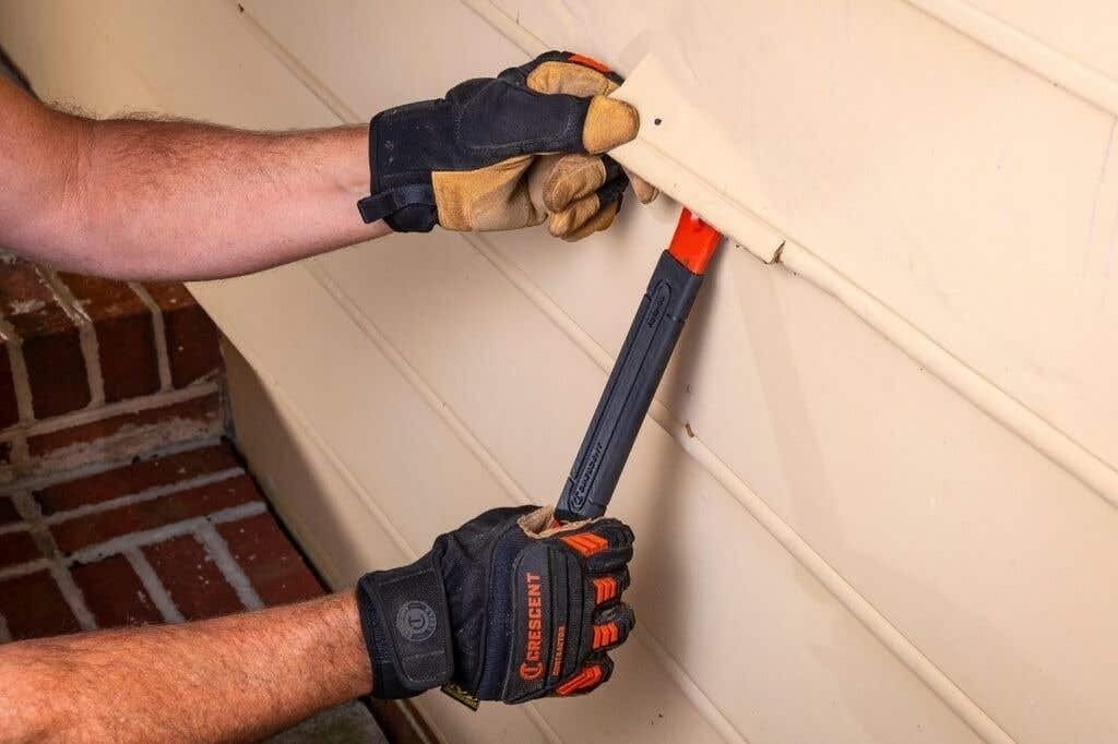 Close-up action shot showing a worker using the pry bar to lift a piece of beige vinyl siding, demonstrating the tool's ability to fit into narrow seams for renovation work.
