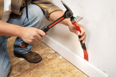 An action photograph of a professional using a Crescent hammer to strike the high-leverage head of the Crescent 15-inch flat pry bar to remove white baseboard trim from a wall during a renovation project.