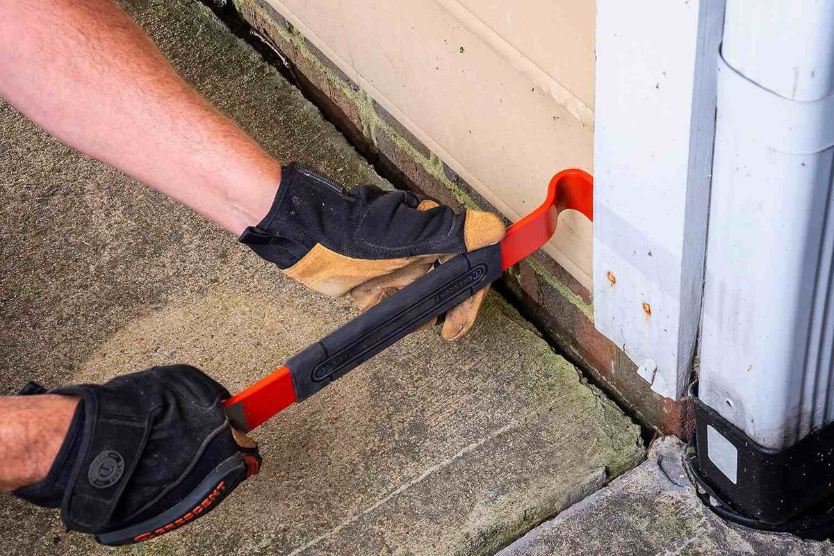 Action shot showing a professional tradesperson using the Crescent flat pry bar to pry a wooden door frame away from a brick exterior, demonstrating the tool's strength and leverage in heavy-duty demolition.