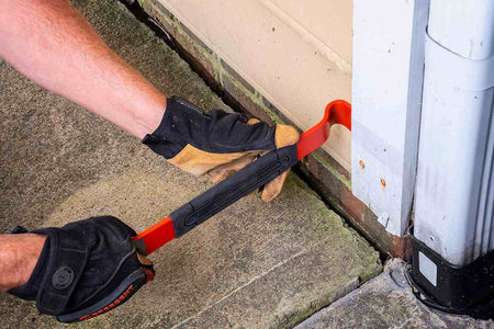 Action shot showing a professional tradesperson using the Crescent flat pry bar to pry a wooden door frame away from a brick exterior, demonstrating the tool's strength and leverage in heavy-duty demolition.