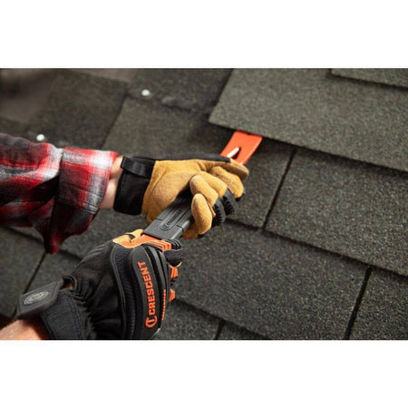 An action photograph of a worker wearing professional gloves using the Crescent flat pry bar to lift and remove asphalt roof shingles, highlighting the versatility of the beveled nail slots for different renovation tasks.