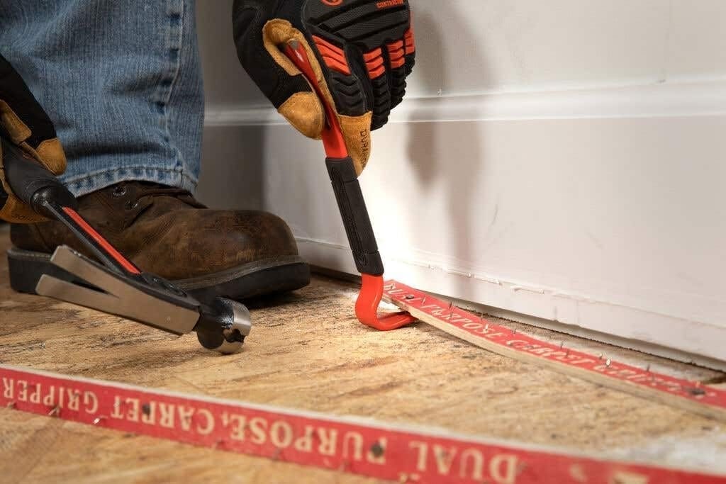 A worker using the gooseneck end of the Crescent 7-inch flat pry bar to carefully lift a carpet tack strip from a wooden subfloor during a home renovation project.