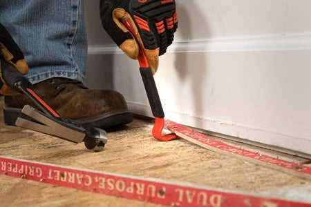 A worker using the gooseneck end of the Crescent 7-inch flat pry bar to carefully lift a carpet tack strip from a wooden subfloor during a home renovation project.
