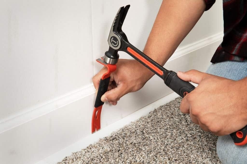 Close-up action shot of a worker using a hammer to strike the integrated striking surface of the Crescent FB7-06 pry bar to drive the tip behind a white baseboard.