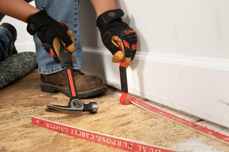 Action shot showing a construction professional using the Crescent comfort grip pry bar and a hammer to remove floor-level carpet grippers from a subfloor.