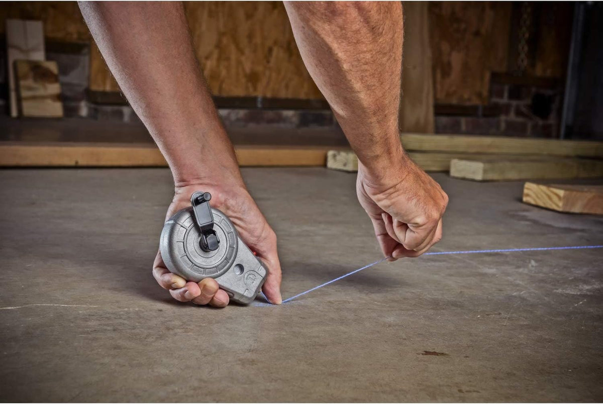 Action shot of a professional technician using the Crescent Lufkin aluminum chalk reel to snap a precise blue layout line on a concrete floor in a workshop environment.