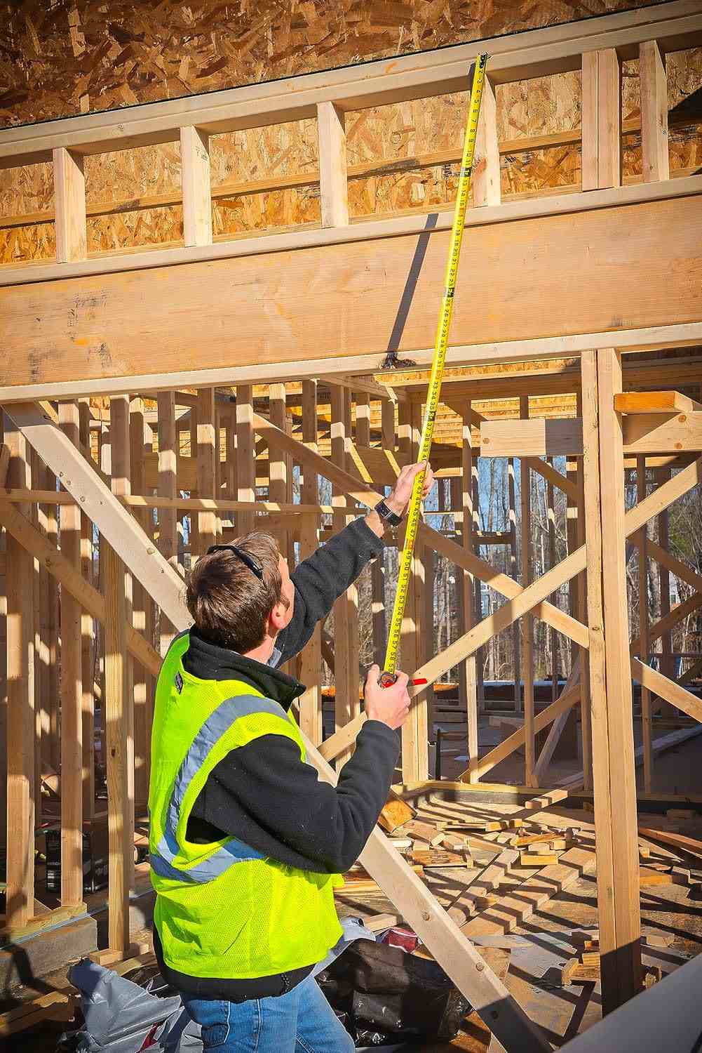 An action photograph showing a professional tradesman in a safety vest using the Crescent Lufkin Shockforce G1 to measure high wooden framing, illustrating the tape's 15-foot reach and solo-measuring efficiency.