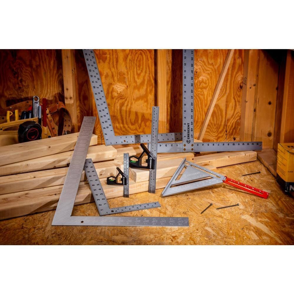 A lifestyle shot showing a variety of Crescent layout tools, including the L1624RS aluminum rafter square and combination squares, arranged on a wooden workbench among lumber and nails to demonstrate professional carpentry utility.