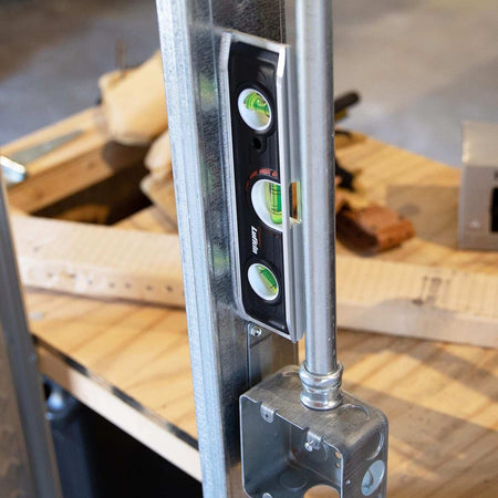 A lifestyle action shot showing the Crescent Lufkin torpedo level magnetically secured to a vertical conduit pipe above an electrical box, highlighting its hands-free capability for electrical and plumbing professional work.