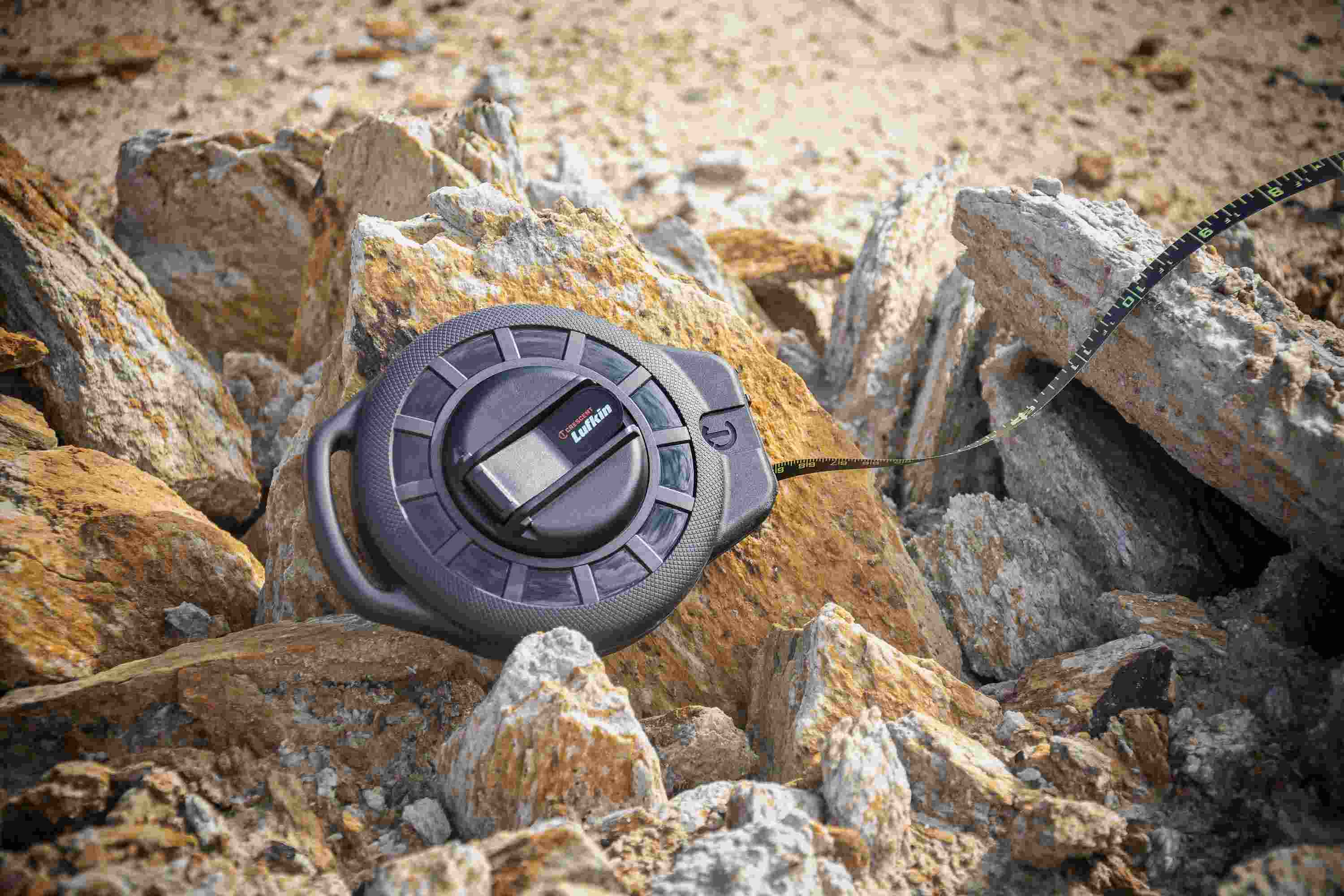 A lifestyle photograph of the Crescent Lufkin ST100B-07 long tape resting on rugged rocks at a construction site with the black steel blade extended, illustrating the tool's durability in harsh outdoor environments.