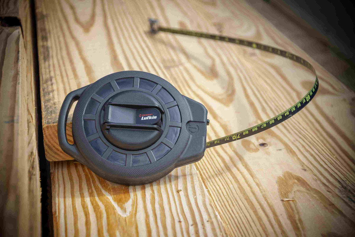 A perspective lifestyle shot of the Crescent Lufkin 100-foot Nite Eye tape measure resting on wooden framing, highlighting the compact ergonomic ABS case and the high-contrast green-on-black measuring scale.
