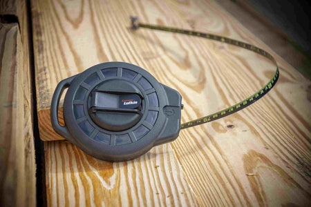 A perspective lifestyle shot of the Crescent Lufkin 100-foot Nite Eye tape measure resting on wooden framing, highlighting the compact ergonomic ABS case and the high-contrast green-on-black measuring scale.