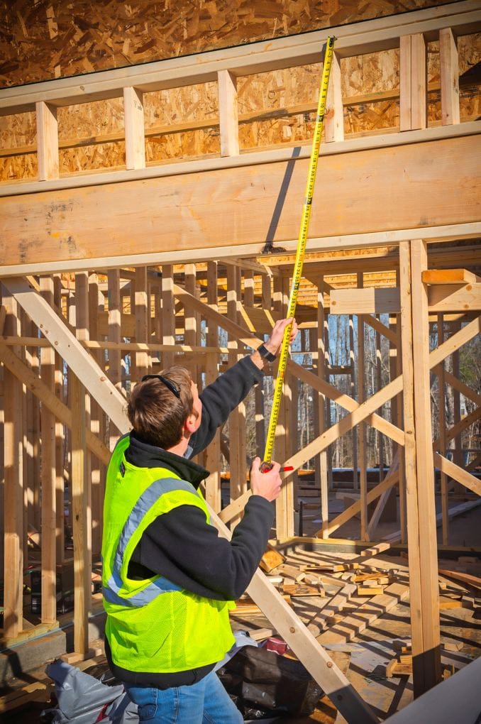 Demonstration of the 14-foot standout capability of the Shockforce G1 tape measure, shown being used vertically by a professional worker to measure a wooden wall frame.