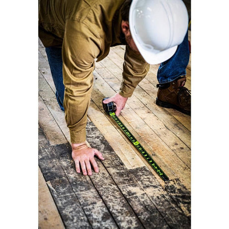 A construction worker kneeling on a wooden floor using the extended high-visibility tape measure to layout measurements.