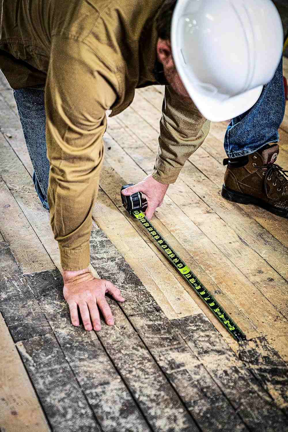 Overhead view of a trade professional wearing a white hard hat using a Crescent Lufkin Shockforce Nite Eye 25-foot tape measure to mark long measurements on a rustic wooden floor.