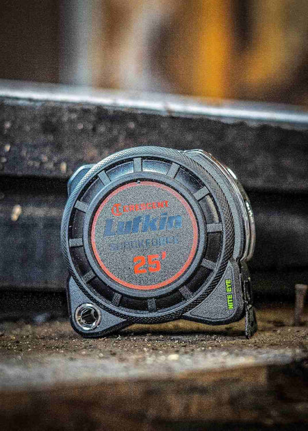 Close-up of the Crescent Lufkin Shockforce Nite Eye 25-foot tape measure case resting on a rugged, dark industrial surface, highlighting the durable impact-resistant housing and Nite Eye branding.