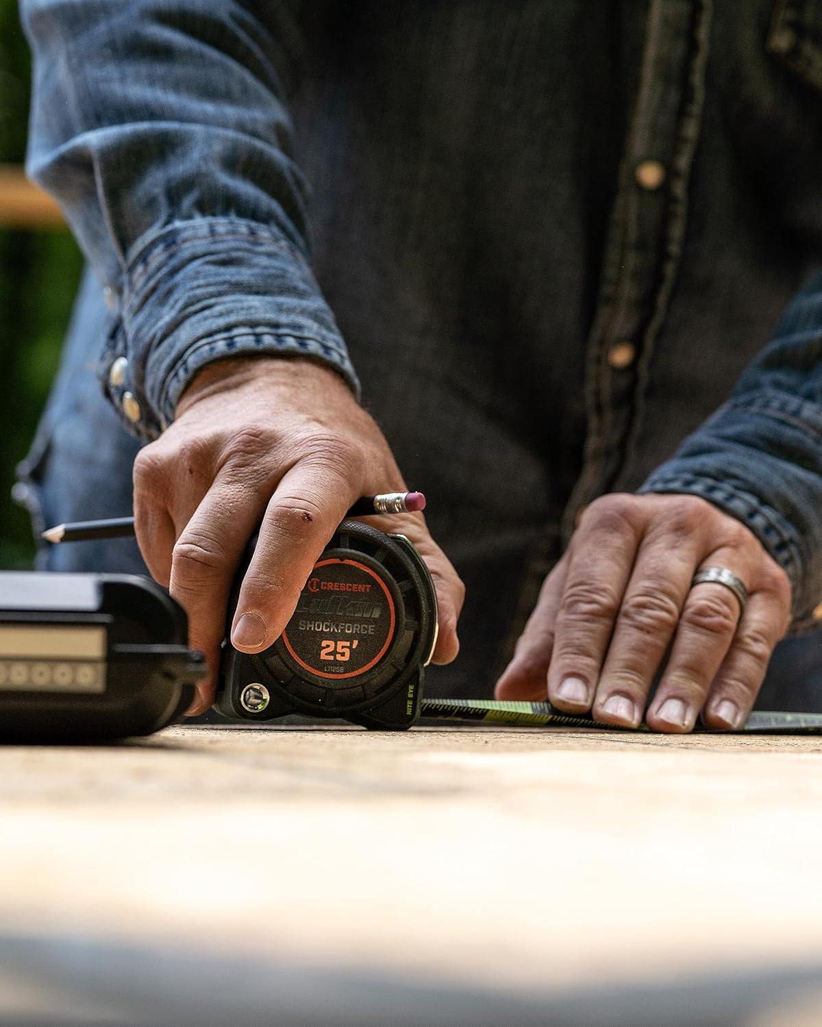 Close-up of a professional's hands using the Crescent Lufkin L1125B-02 tape measure to mark a measurement on a wooden surface with a pencil, showcasing the compact case design.