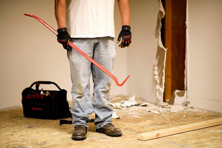 A worker holding the 42-inch Crescent wrecking bar on a jobsite next to a tool bag, illustrating the tool's large scale and suitability for heavy demolition tasks.