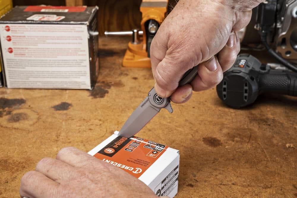 A close-up action view of a user using the fine edge of the Crescent folding pocket knife to slice open a cardboard retail box of Crescent tools on a wooden workshop table.