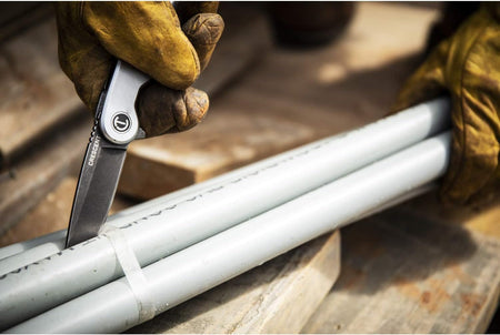 A vertical perspective action photograph of a worker in leather safety gloves guiding the stonewashed D2 tool steel blade of the Crescent folding utility knife to cut through heavy industrial tape securing bundles of grey PVC construction pipes.
