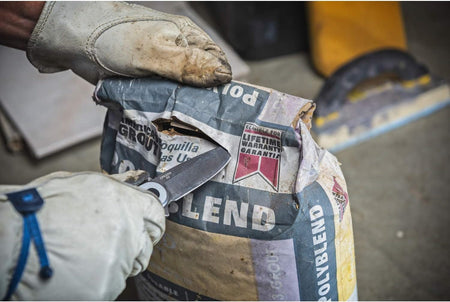 A worker wearing thick leather safety gloves utilizes the stonewashed D2 steel blade of the Crescent CPK325A folding pocket utility knife to slice open a heavy paper bag of Polyblend commercial grout on a jobsite.