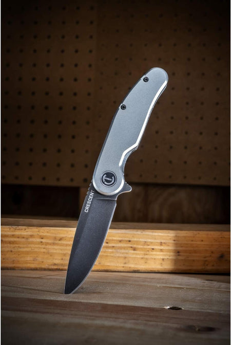 The Crescent folding pocket utility knife standing vertically with its 3-1/4 inch drop point blade embedded into a wooden work surface, showcasing the silver anodized aluminum handle scale design against a workshop background.
