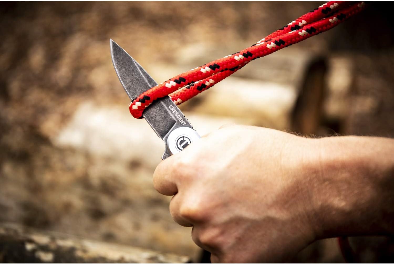 Action closeup of a user holding the Crescent pocket utility knife by its silver handle, utilizing the sharp drop point tip of the D2 steel blade to score and slice down through a thick red, black, and white braided nylon utility rope.