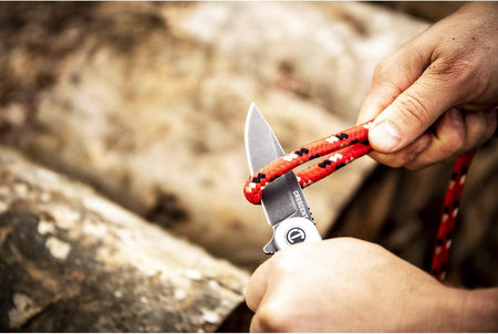 An alternate macro action shot of the manual folding utility knife slicing cleanly through a braided red utility cord, demonstrating the high-performance structural cutting leverage and finger contact safety ergonomics of the handle design.