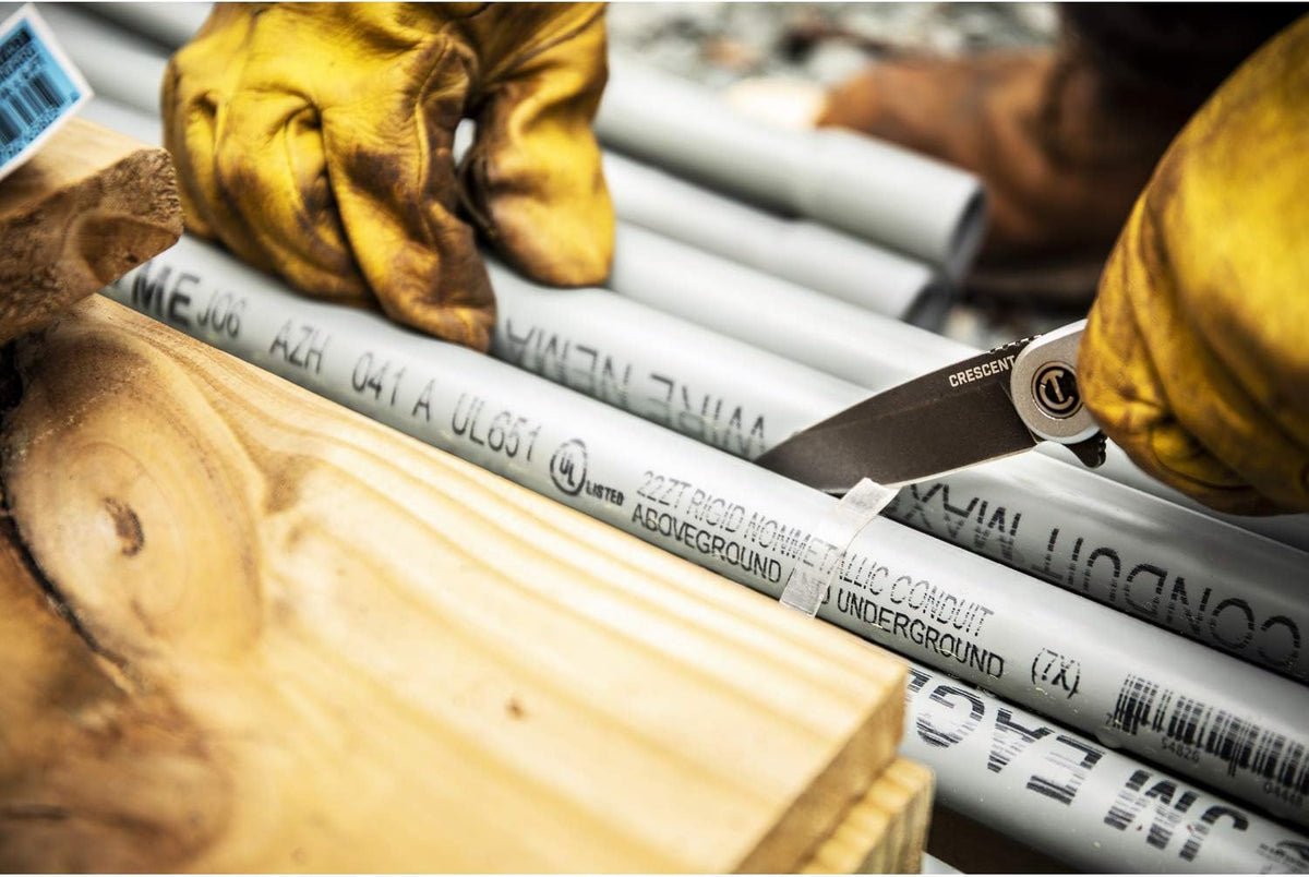 Jobsite action view of a tradesman wearing yellow leather work gloves using the Crescent CPK325A pocket knife to strip heavy plastic wrapping material from bundled bundles of gray rigid non-metallic electrical conduit pipes.