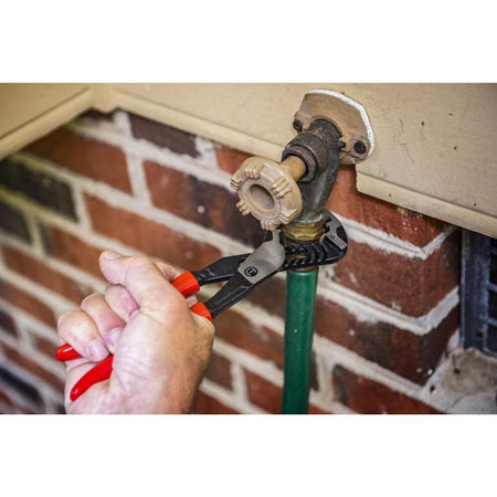 An action photo of a plumber using the Crescent RTZ210 straight jaw slip joint pliers to tightly grip and turn a brass plumbing fitting on an outdoor water spigot and green garden hose against a red brick wall.