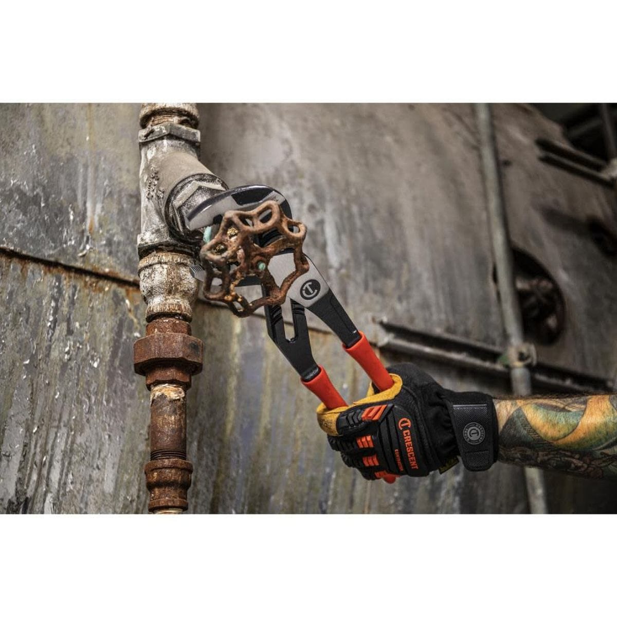 A technician wearing heavy duty Crescent branded work gloves using the 10 inch adjustable water pump pliers to grip and turn a large rusted metal pipe coupling on an industrial machinery line.
