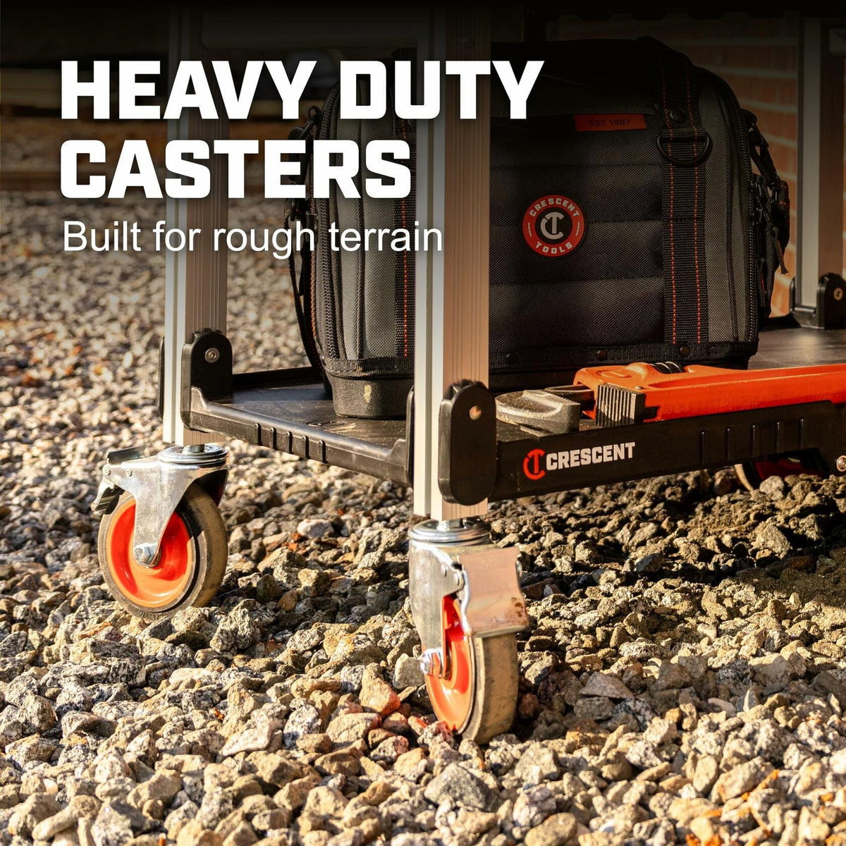 Close-up of Crescent jobsite cart wheels on rough gravel terrain. The cart features bright orange heavy-duty casters and a black tool bag with red accents. A red Crescent tool is also visible. Text overlay reads “HEAVY DUTY CASTERS – Built for rough terrain.”