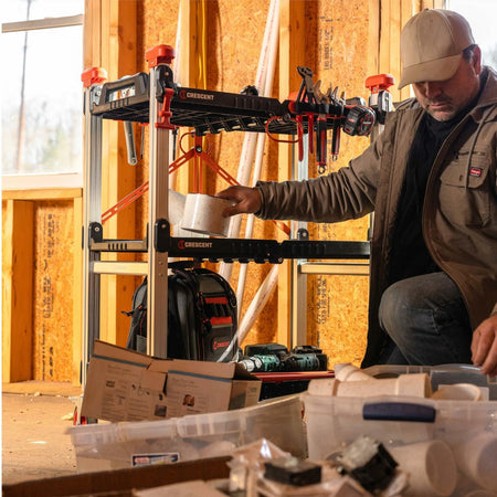 Man kneeling beside a Crescent-branded jobsite cart in a construction setting. The cart holds hand tools, a black and red Crescent backpack, and power tools across multiple shelves. The background shows unfinished wooden walls with insulation, and open bins with documents and materials are visible in the foreground.