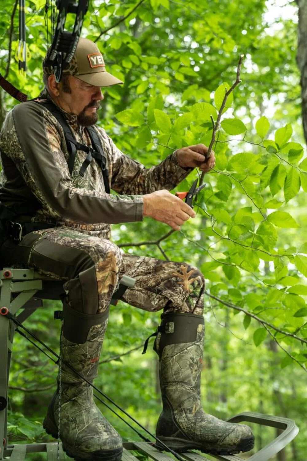 A full-body lifestyle shot of a hunter or tradesman in camouflage gear using the Crescent Wiss tradesman utility shears to clear small brush, demonstrating real-world versatility for heavy-duty outdoor and job site tasks.