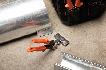 A lifestyle photograph showing the Crescent Wiss WS4N offset seamer resting on a concrete floor next to galvanized sheet metal ducting and a professional tool bag, highlighting its use in HVAC installations.
