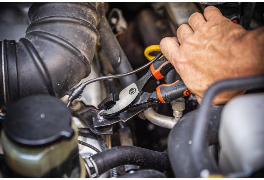 Action shot of a hand using the Crescent Z2 slip joint pliers to grip a metal bracket fastener inside a crowded vehicle engine bay.