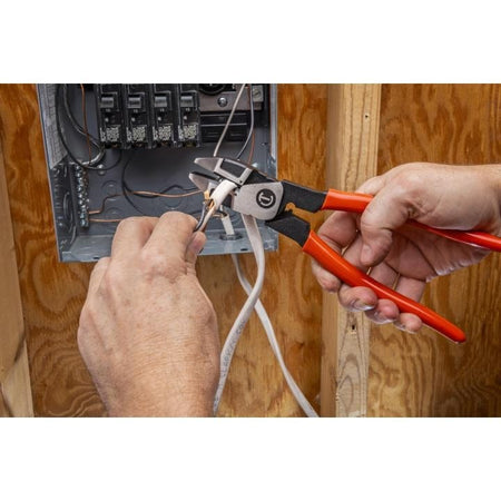 Close-up of a person using red-handled diagonal cutting pliers to cut copper wire inside an electrical panel. Circuit breakers and connected wires are visible, with wooden framing in the background, showing a typical electrical installation task