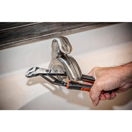 A real-world plumbing application photograph showing a tradesman using the Crescent 6-inch quick-adjust V-jaw pliers to tighten a connection behind a brushed nickel bathroom sink faucet.