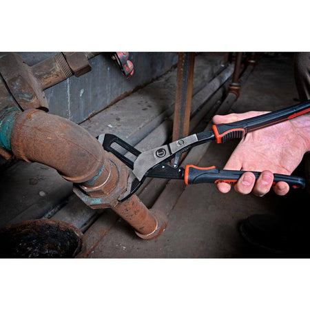 An industrial facility maintenance photo showing a plumber using the drop-forged alloy steel jaws of the Crescent 6-inch tongue and groove pliers to grip and turn a heavily rusted iron pipe coupling in a crawlspace.