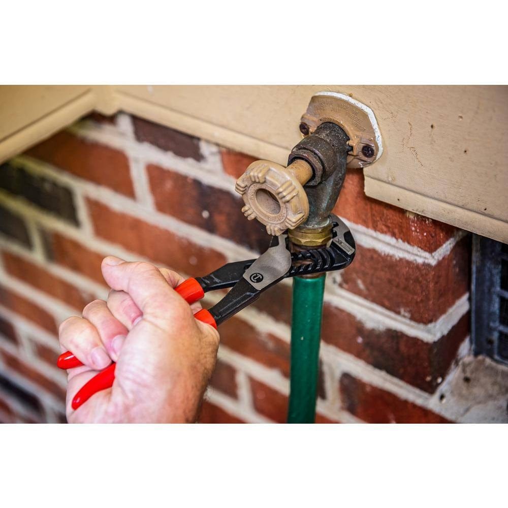 Close-up of red-handled adjustable pliers gripping a brass outdoor faucet connected to a green garden hose. The faucet is mounted on a beige panel above a brick wall, showing outdoor plumbing maintenance.