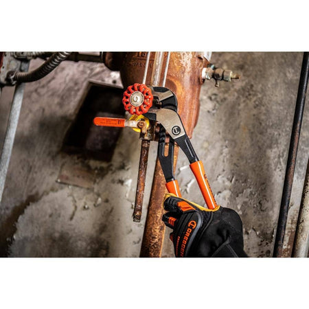 Person using Crescent tongue and groove pliers with orange handles to turn a red valve on a rusted pipe. The setting includes a concrete wall and industrial plumbing, illustrating tool use in maintenance or repair work.