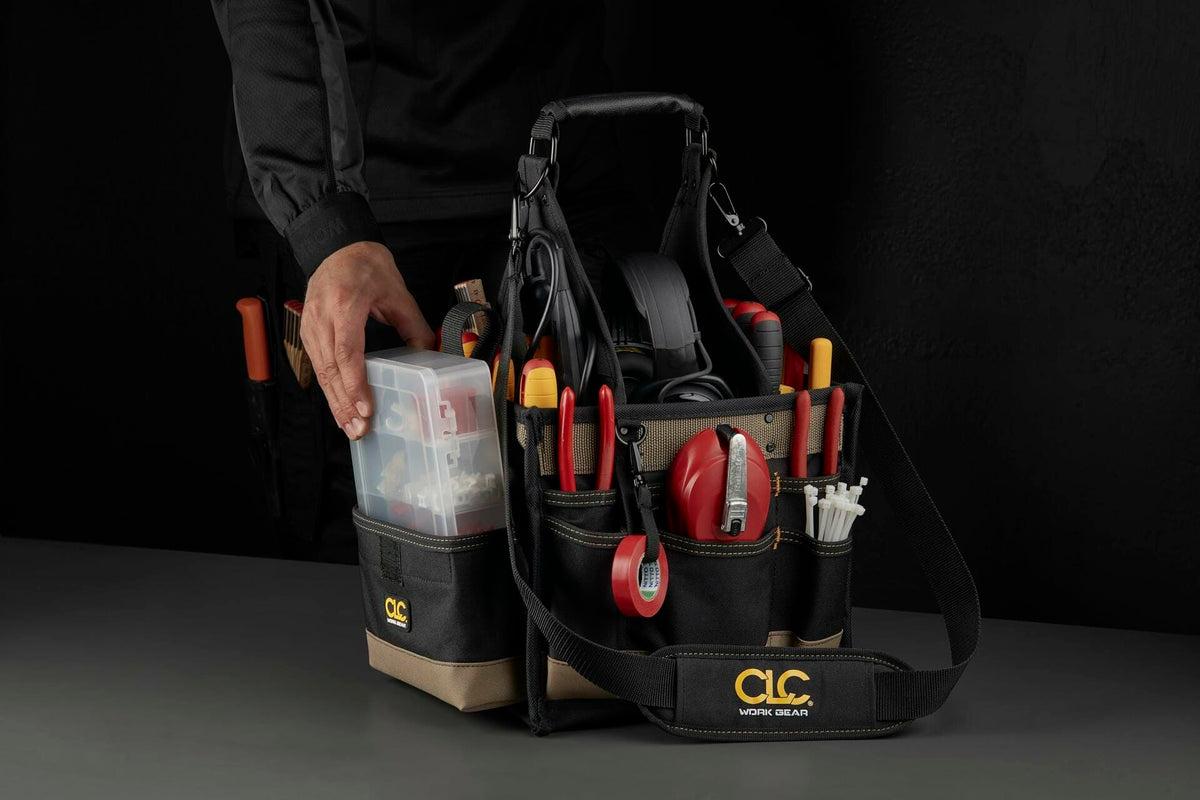 A worker's hand reaching to place the clear plastic parts tray into a pocket of the loaded CLC tool carrier bag, set against a dark background.