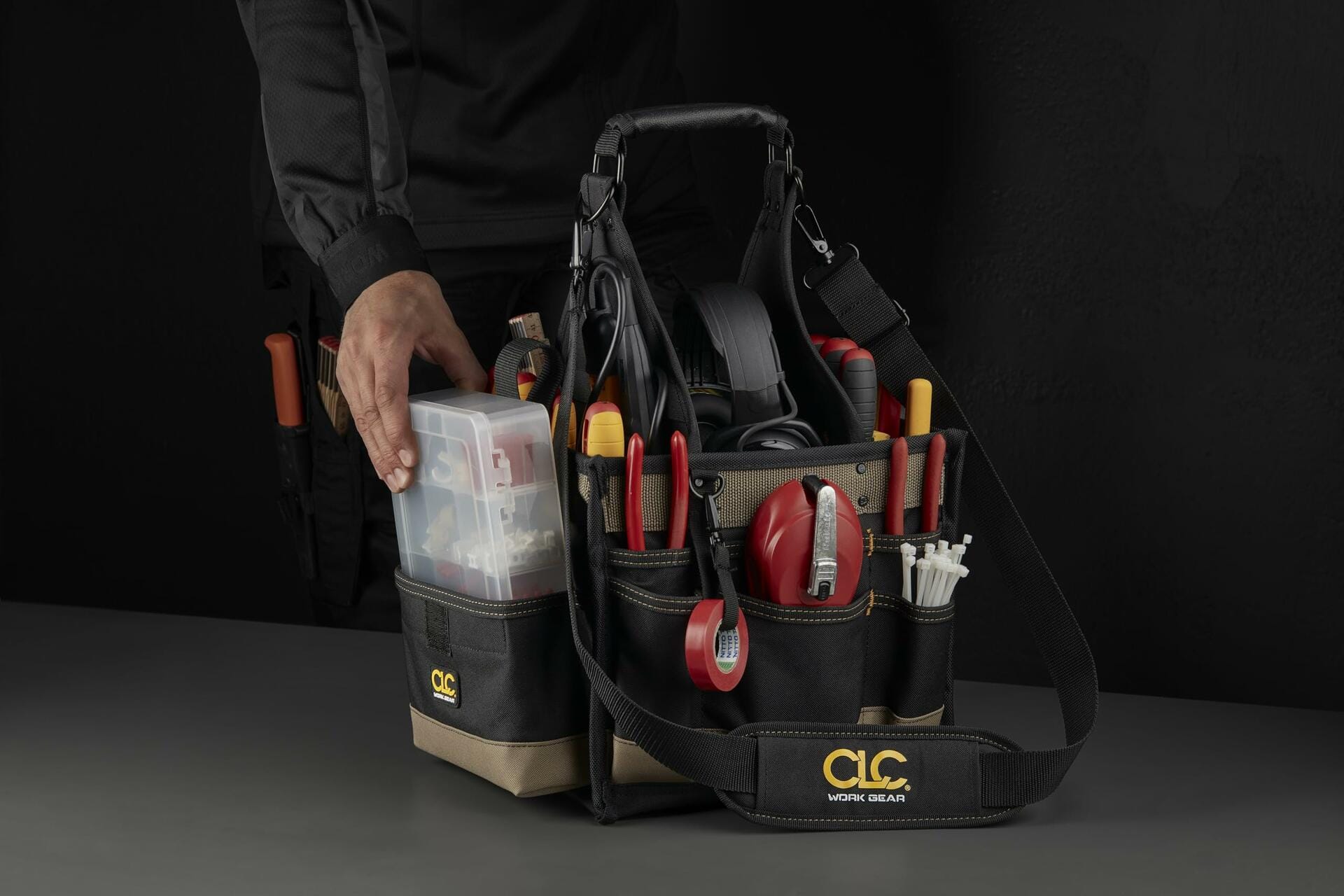 A worker's hand reaching to place the clear plastic parts tray into a pocket of the loaded CLC tool carrier bag, set against a dark background.