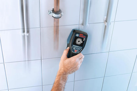 Hand holding Bosch D-TECT 120 scanner against tiled wall; display shows detection of metal pipe behind surface.