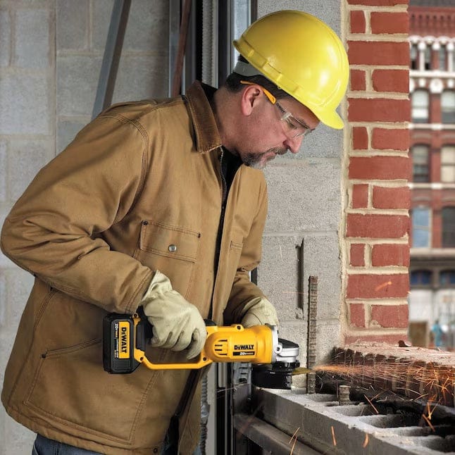 Construction worker wearing safety gear using a DEWALT cordless angle grinder to grind metal at a jobsite; sparks visible from grinding point.