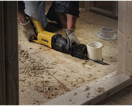 DEWALT reciprocating saw cutting through a white PVC pipe on a wooden subfloor, with wood shavings and exposed framing visible in a renovation setting.