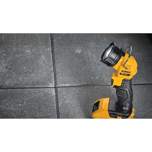 A DEWALT DCL040 LED work light stands upright on a construction site floor, illuminating an electrical box while a gloved worker uses a wire stripper tool. The scene highlights hands-free lighting during installation.
